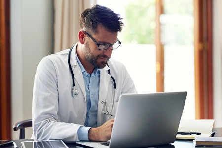 male doctor sitting at office desk typing on laptop