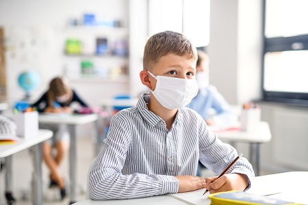 young boy wearing face mask sitting at desk in classroom