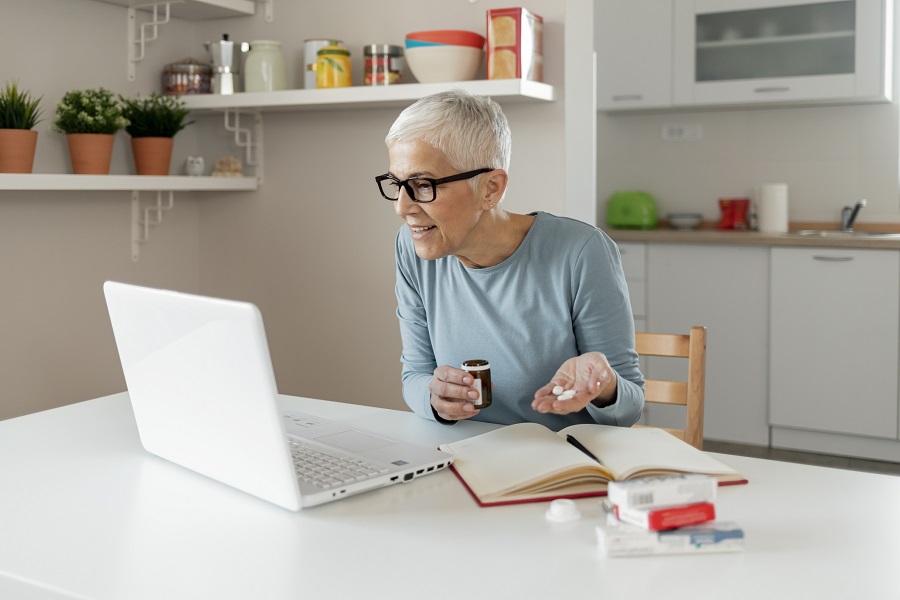 Une femme âgée tenant des pilules et un flacon de médicament lit un écran d'ordinateur portable
